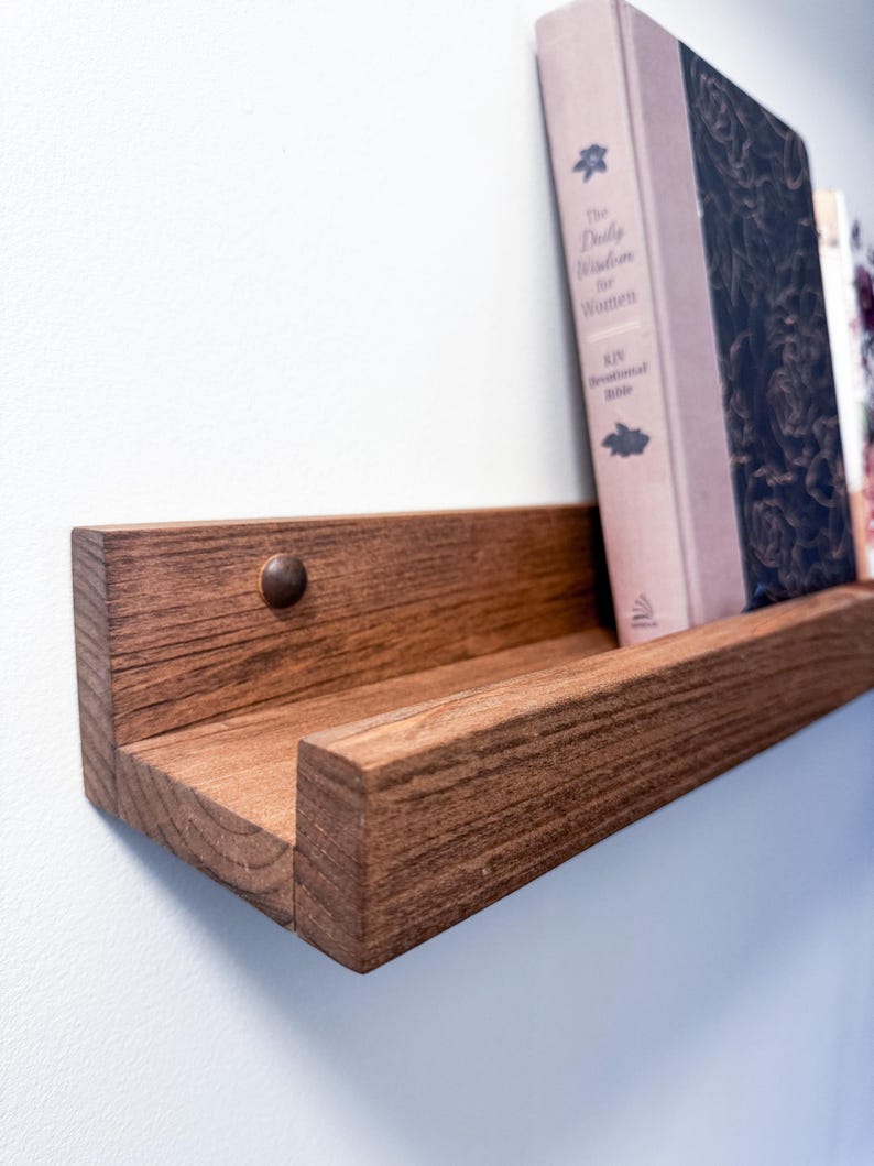 May include: A dark brown wooden shelf with a book titled "The Daily Wisdom for Women" resting on it.