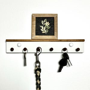 May include: White key rack with five hooks and brown wooden shelf. The rack has a small framed print of a green plant on the shelf. There are keys and a leash hanging on the hooks.