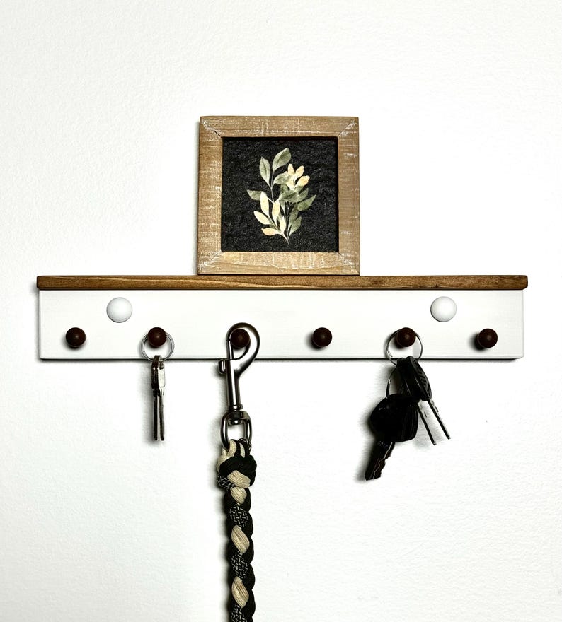 May include: White key rack with five hooks and brown wooden shelf. The rack has a small framed print of a green plant on the shelf. There are keys and a leash hanging on the hooks.