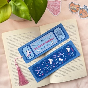 Op de afbeelding: Twee blauwe bladwijzers met de tekst "The Ice Hockey Romance Book Club" en illustraties van schaatsen en hockeysticks. Een roze kwast hangt aan een bladwijzer. De bladwijzers liggen op een open boek.