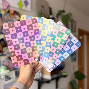 May include: A hand displays a selection of holographic cards. Each card has a checkered design with star-shaped details. The cards show a spectrum of colours, including pink, yellow, green, and blue, with a shimmering, iridescent finish.
