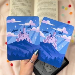 Könnte beinhalten: Zwei blaue Fantasy-Kunstdrucke mit einem Schloss auf einem Berg, Drachen, Wolken und Sternen. Die Drucke werden über einem offenen Buch und einem Kindle-E-Reader gehalten. Die Drucke haben einen hellblauen Himmel und einen dunkelblauen Berg.