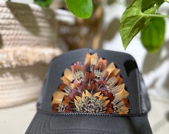 Pode incluir: Boné de caminhoneiro cinza carvão com um arranjo decorativo de penas marrons, bege e douradas no painel frontal. O boné tem uma parte traseira de malha e uma aba curva, oferecendo um acessório elegante.