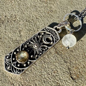 Può includere: Una collana d'argento con un ciondolo rettangolare che presenta un design di sole, luna e fiori. Il ciondolo ha una pietra preziosa trasparente al centro. La collana ha anche un piccolo ciondolo di cristallo trasparente.
