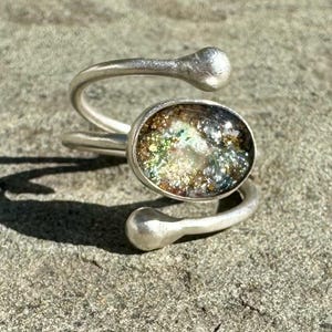 Può includere: Un anello d'argento con un design a spirale e una pietra rotonda in vetro iridescente incastonata al centro. La pietra ha una miscela di colori dorati, verdi e blu.