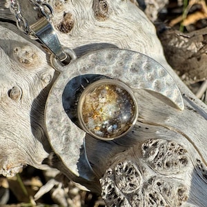 Puede incluir: Un collar con un colgante de media luna de plata con un dije redondo de resina transparente lleno de purpurina dorada y blanca.