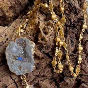 Peut inclure: Un collier en chaîne dorée avec un grand pendentif en agate blanc iridescent et une petite goutte de quartz rose. Le collier est présenté sur un morceau de bois usé.