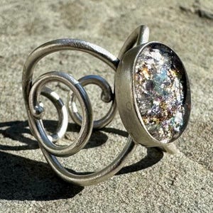 Könnte beinhalten: Silbernes Armreif-Armband mit einem geschwungenen Design und einem großen ovalen Harz-Cabochon mit eingebetteten schillernden Glitzer.