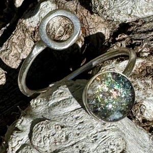 Könnte beinhalten: Ein silberner Ring mit einem runden, offenen Design und einem kreisförmigen, schillernden Glasstein.