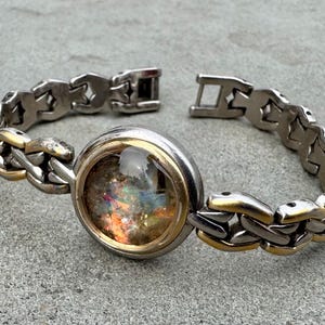 May include: Silver-tone bracelet with a large, round, iridescent center stone. The bracelet features a chain-link design with gold-tone accents. The clasp is open, showing the bracelet's construction. The stone has a mix of colors.