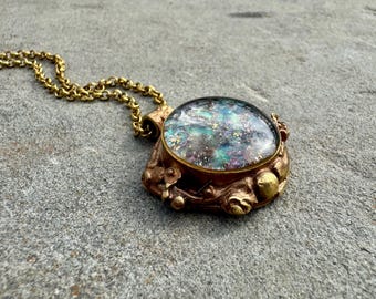 May include: A gold necklace with a round pendant featuring a glittery, iridescent resin design. The pendant is surrounded by a detailed, textured metal frame.