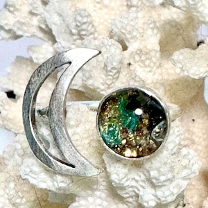 Peut inclure: Une bague en argent composée d'un croissant de lune et d'une pierre ronde sertie. La pierre présente des couleurs vertes, dorées et marron. La bague est présentée sur une surface blanche texturée.