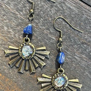 May include: A pair of gold tone earrings with a sunburst design and a blue stone dangle. The earrings have a hook closure.