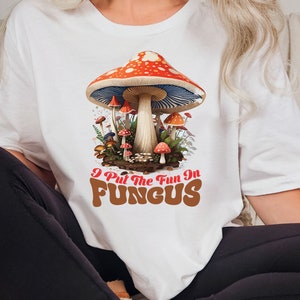 May include: White t-shirt with a graphic of red and white mushrooms with a blue cap. The text "I Put The Fun In FUNGUS" is printed in brown letters with a retro font.