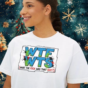 May include: White t-shirt with the text "WTF WTS" in blue and green, with red and black paint splatters. The phrase "(WHAT THE FUCK WAS THAT SHIT?)" is below the main text. The shirt is being worn by a person.