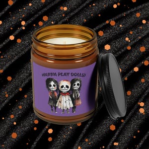 Peut inclure: Un bocal en verre brun contenant une bougie avec une étiquette violette sur laquelle est inscrit "Wanna Play Dolls?" et une image de trois poupées effrayantes. Le couvercle est noir.