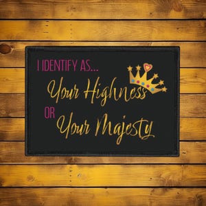 May include: Black rectangular patch with the text "I IDENTIFY AS... Your Highness OR Your Majesty" in gold script and pink block letters. A gold crown with a pink heart is in the upper right corner.