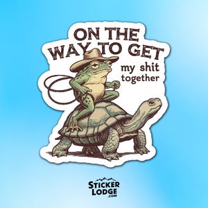 May include: A sticker featuring a cartoon frog wearing a cowboy hat riding a turtle. The text reads "ON THE WAY TO GET my shit together". The sticker has a white border and a blue background.
