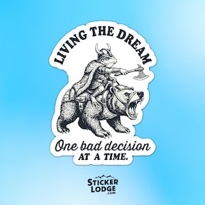 May include: A black and white sticker featuring a frog in a Viking helmet riding a bear and holding an axe. The text "LIVING THE DREAM" arches above the frog and bear. Below, the text reads "One bad decision at a time."