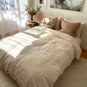 May include: A bedroom scene featuring a bed with a cream-colored duvet cover and pillows. Decorative pillows with floral and plaid patterns add visual interest. A vase of flowers sits on a small table next to the bed. A large painting and framed art pieces adorn the wall.
