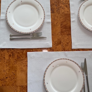 May include: Four place settings featuring white plates with a decorative border of blue, red, and yellow floral accents. Each plate sits on a white chevron-patterned placemat, with a fork and knife.