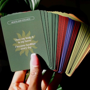 May include: A deck of rectangular affirmation cards fanned out, featuring a dark green top card with the text "HEALTH AND WELLNESS" and "I love my body, it is my home. I become healthier every day" printed over a yellow sun graphic.