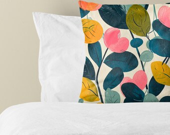 Puede incluir: Un cojín cuadrado con fondo color crema y un patrón botánico de hojas azul verdoso oscuro y flores redondeadas en tonos rosa brillante, naranja y amarillo. El diseño presenta una textura pintada con pinceladas visibles.