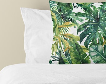 Puede incluir: Un cojín cuadrado con un estampado botánico vibrante de grandes hojas verdes de monstera y palmera sobre un fondo blanco. El cojín está colocado en posición vertical contra una funda de almohada y un edredón blancos.