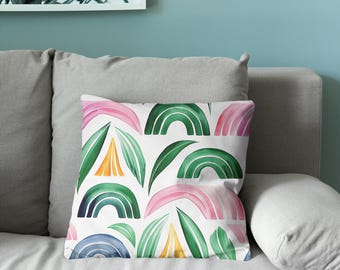 Puede incluir: Un cojín cuadrado con fondo blanco y un patrón repetitivo de formas abstractas. El diseño incluye arcos similares a arcoíris en tonos rosa, verde y azul, intercalados con formas puntiagudas que recuerdan a hojas en colores verde y amarillo.
