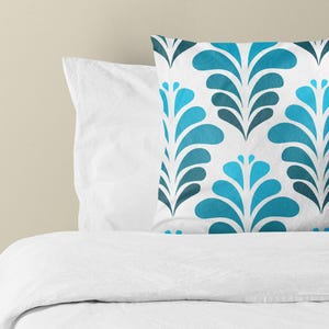 May include: A square throw pillow featuring a repeating pattern of stylized, symmetrical leaf-like shapes in shades of bright blue, teal, and dark navy blue against a crisp white background, positioned atop a plain white bedspread.