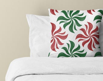 Puede incluir: Un cojín cuadrado con un patrón repetitivo de formas giratorias tipo molinete en colores verde oscuro y rojo intenso que se alternan sobre un fondo blanco, colocado sobre una cama con ropa de cama blanca.