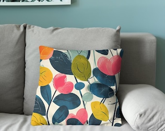 Puede incluir: Un cojín cuadrado con un estampado botánico sobre un fondo crema. El diseño incluye hojas estilizadas y formas redondeadas en tonos azul oscuro, amarillo brillante, naranja, verde oliva y rosa, dispuestos en una composición densa y superpuesta.