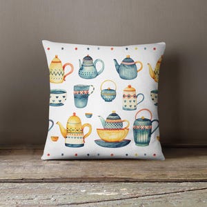 May include: A square throw pillow featuring a white background with a repeating watercolor pattern of various teapots, teacups, and bowls in shades of blue, yellow, and red, accented by a border of small colored dots along the top and bottom.