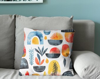 Puede incluir: Un cojín decorativo con fondo blanco y un patrón de acuarela. El patrón incluye formas abstractas en azul, naranja, amarillo y negro. El cojín está sobre un sofá gris.