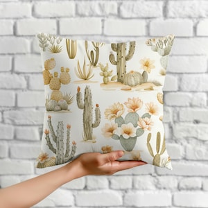 May include: A square throw pillow featuring a repeating pattern of various green and tan cacti and succulents on a white background, including tall saguaro-style cacti, prickly pear cacti, and several blooming flowers in shades of pale yellow.
