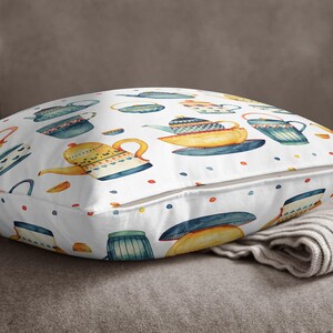 May include: A square throw pillow featuring a white background with a repeating watercolor pattern of various teapots and teacups in shades of yellow, blue, and orange, accented by small colorful dots scattered across the fabric surface.