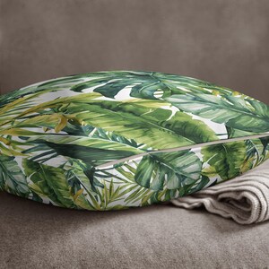 May include: A square throw pillow featuring a vibrant botanical print of overlapping tropical palm and monstera leaves in various shades of green, yellow, and white. The fabric has a smooth texture and a visible seam along the edge.
