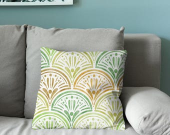 Puede incluir: Un cojín cuadrado con un patrón repetitivo en forma de abanico en tonos acuarela verde y dorado. El diseño consiste en semicírculos superpuestos con líneas verticales y pequeños puntos, creando un motivo decorativo simétrico.