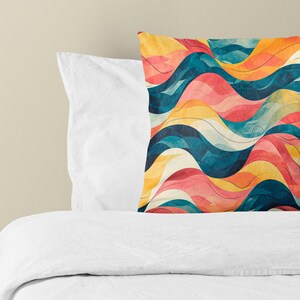 May include: A square throw pillow featuring a repeating pattern of horizontal wavy lines in shades of deep teal, vibrant orange, soft pink, and cream. The pillow is positioned in front of a plain white pillow and a white bed sheet.