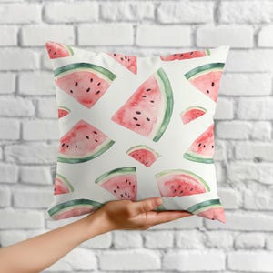 May include: White square throw pillow featuring a watercolor pattern of watermelon slices. The slices are pink with black seeds and green rinds. The pillow is held up by a hand.
