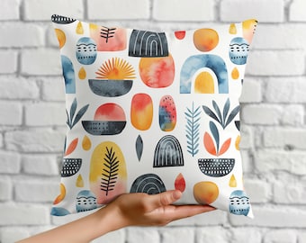 Puede incluir: Un cojín decorativo cuadrado con un patrón de acuarela repetido sobre un fondo blanco. El patrón presenta formas abstractas en tonos de azul, naranja, amarillo y negro. El cojín parece estar hecho de una tela suave.