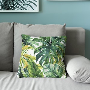 May include: A square throw pillow featuring a vibrant watercolor print of lush green monstera leaves and tropical foliage against a crisp white background. The botanical pattern displays various shades of deep forest green, lime, and yellow-green.