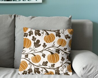 Puede incluir: Un cojín cuadrado con un patrón repetitivo de calabazas naranjas y enredaderas de hojas de color marrón oscuro sobre un fondo blanco. El diseño incluye formas de calabaza estilizadas con tallos visibles y zarcillos.