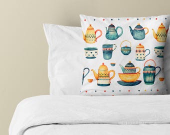 Puede incluir: Un cojín cuadrado con fondo blanco y un patrón de acuarela repetitivo de varias teteras, tazas y cuencos en tonos azul, amarillo y naranja, acentuado por un borde de pequeños puntos de colores.