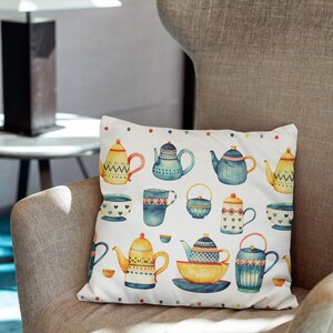 May include: A square throw pillow with a white background featuring a repeating watercolor pattern of various teapots, cups, and bowls in shades of blue, yellow, and orange, accented with small colorful dots along the top and bottom edges.