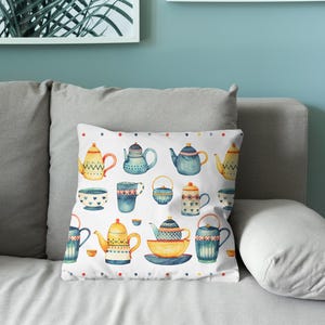 May include: A square throw pillow featuring a white background with a repeating pattern of various teapots, teacups, and bowls in shades of blue, yellow, and orange. The design includes small colorful dots along the top and bottom edges.