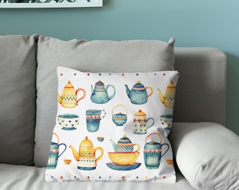 Puede incluir: Un cojín cuadrado con fondo blanco y un patrón repetitivo de varias teteras, tazas de té y cuencos en tonos azules, amarillos y naranjas. El diseño incluye pequeños puntos de colores a lo largo de los bordes superior e inferior.
