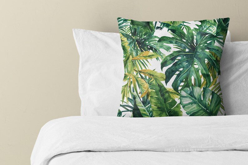 May include: A square throw pillow featuring a vibrant botanical print of large green monstera and palm leaves on a white background. The pillow is positioned upright against a plain white pillowcase and a white duvet cover.