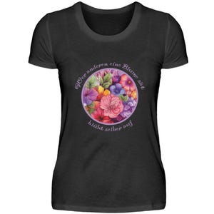 以下が含まれることがあります： カラフルな花束と「Wer anderen eine Blume sät, blüht selber auf」というテキストが書かれた紫色の円が描かれた黒いTシャツです。