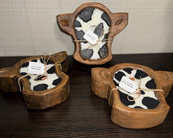 Op de afbeelding: Drie houten kaarsenhouders in de vorm van een koe met zwarte en witte koe-printkaarsen erin. Elke kaarsenhouder heeft een label met de tekst "Small Batch Soy Candles" eraan.